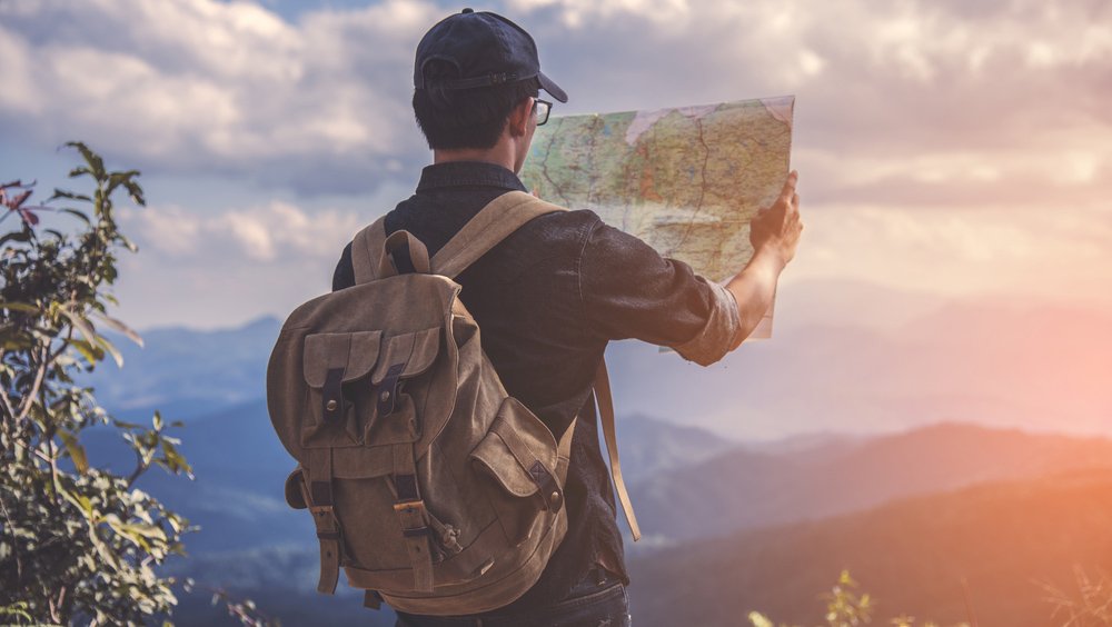 The back of a man wearing a backpack, holding up a map as he's facing the lookout towards rolling mountains