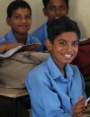 Young male kid sits at a desk smiling with two other young boys sitting behind him