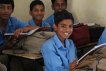 Young male kid sits at a desk smiling with two other young boys sitting behind him